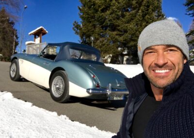 Man standing in front of a classic Austin Healey 3000 on a hill in the snow