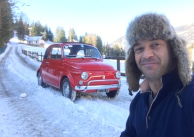 Man smiling in snow in front of a classic red Fiat 500