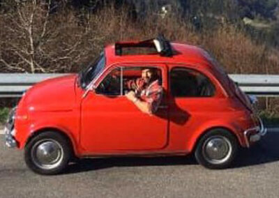 Man in flannel short leaning out of a classic red Fiat 500 on a mountainous road