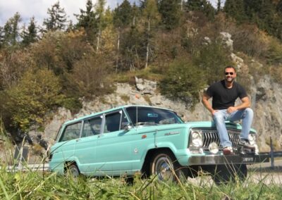 Man sitting on bonnet of turquoise Jeep Kaiser Wagoneer in mountains