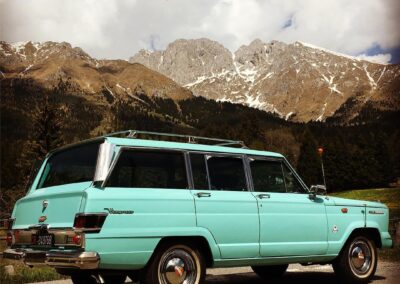 Turquoise vintage Jeep Kaiser Wagoneer parked in front of snow-covered Alps