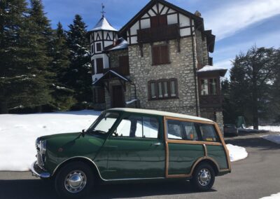 A green classic Mini Traveller parked in the snow in front of a grand house