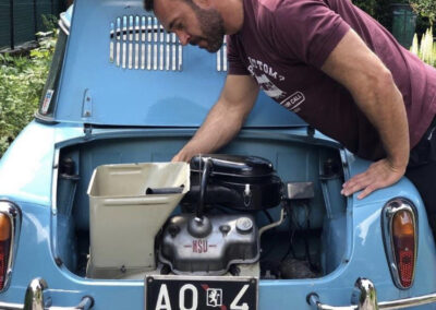 Man leaning into the trunk of a baby blue classic NSU Prinz car, with engine visible