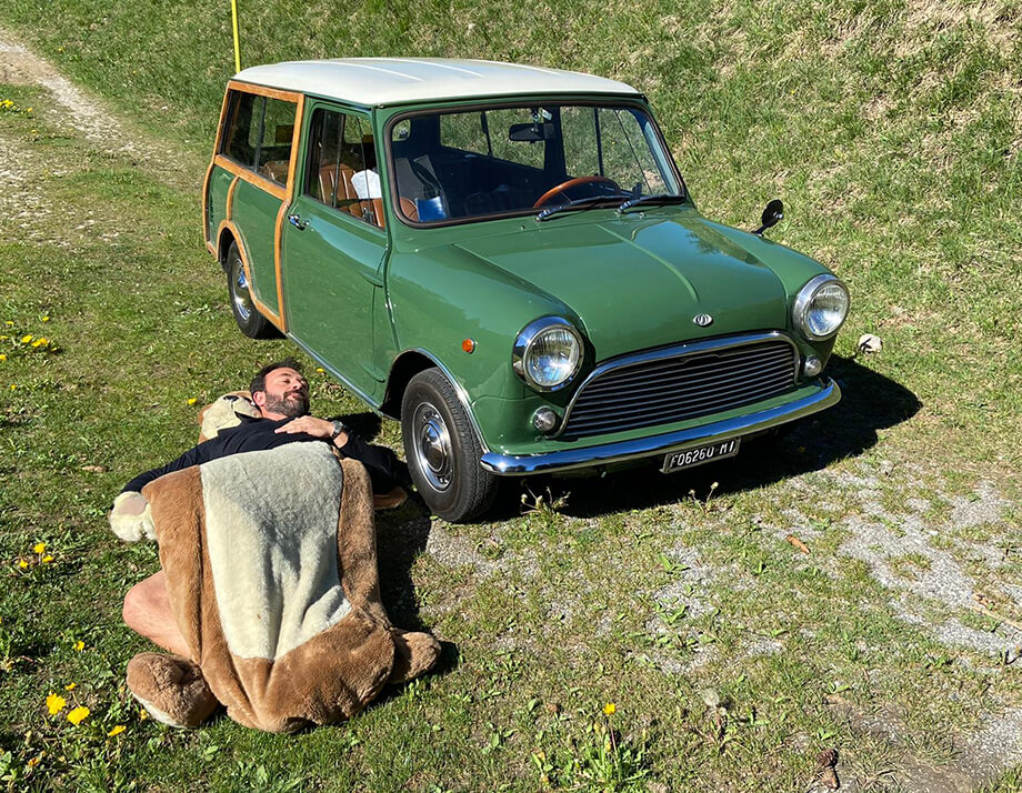 Man sleeping in a field under bear blanket beside a green classic Mini Traveller