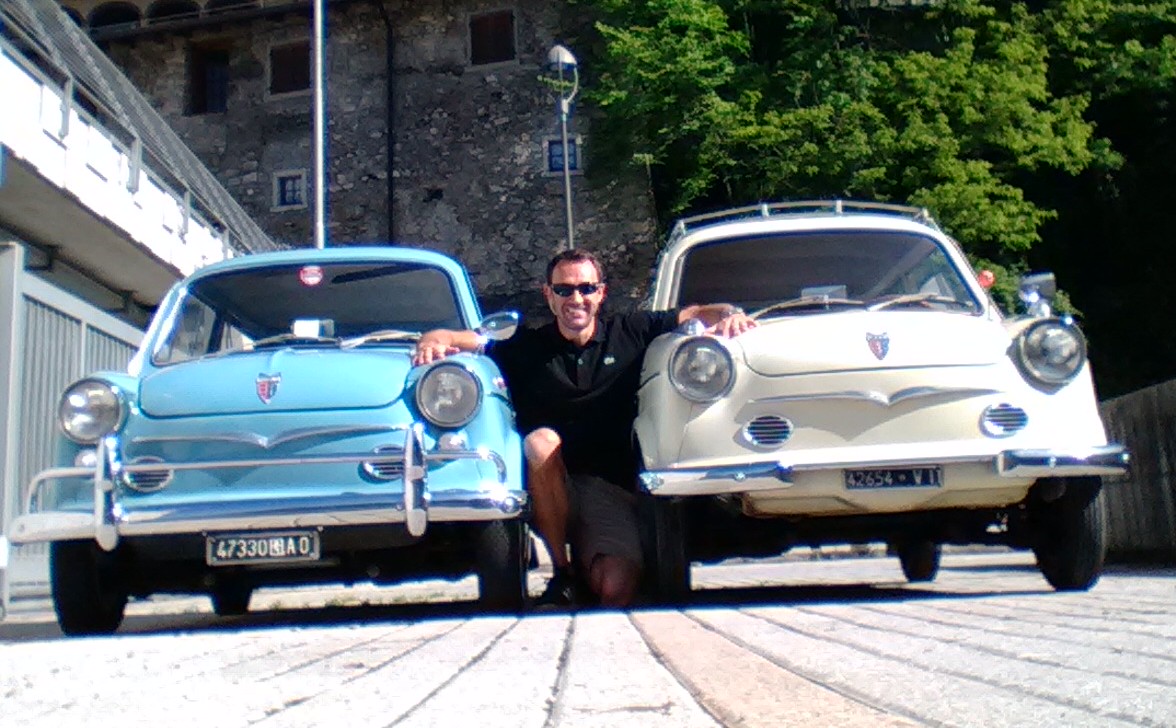 Man leaning down between two classic cars - a baby blue and a beige NSU Prinz
