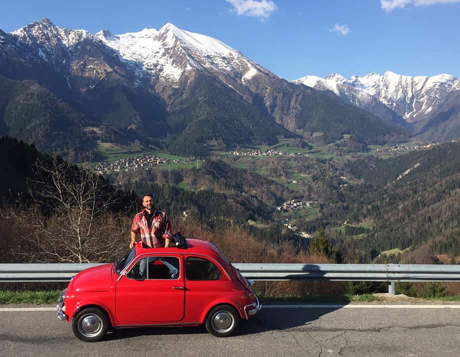 Man standing up through sunroof of a red Fiat 500 on a mountain road